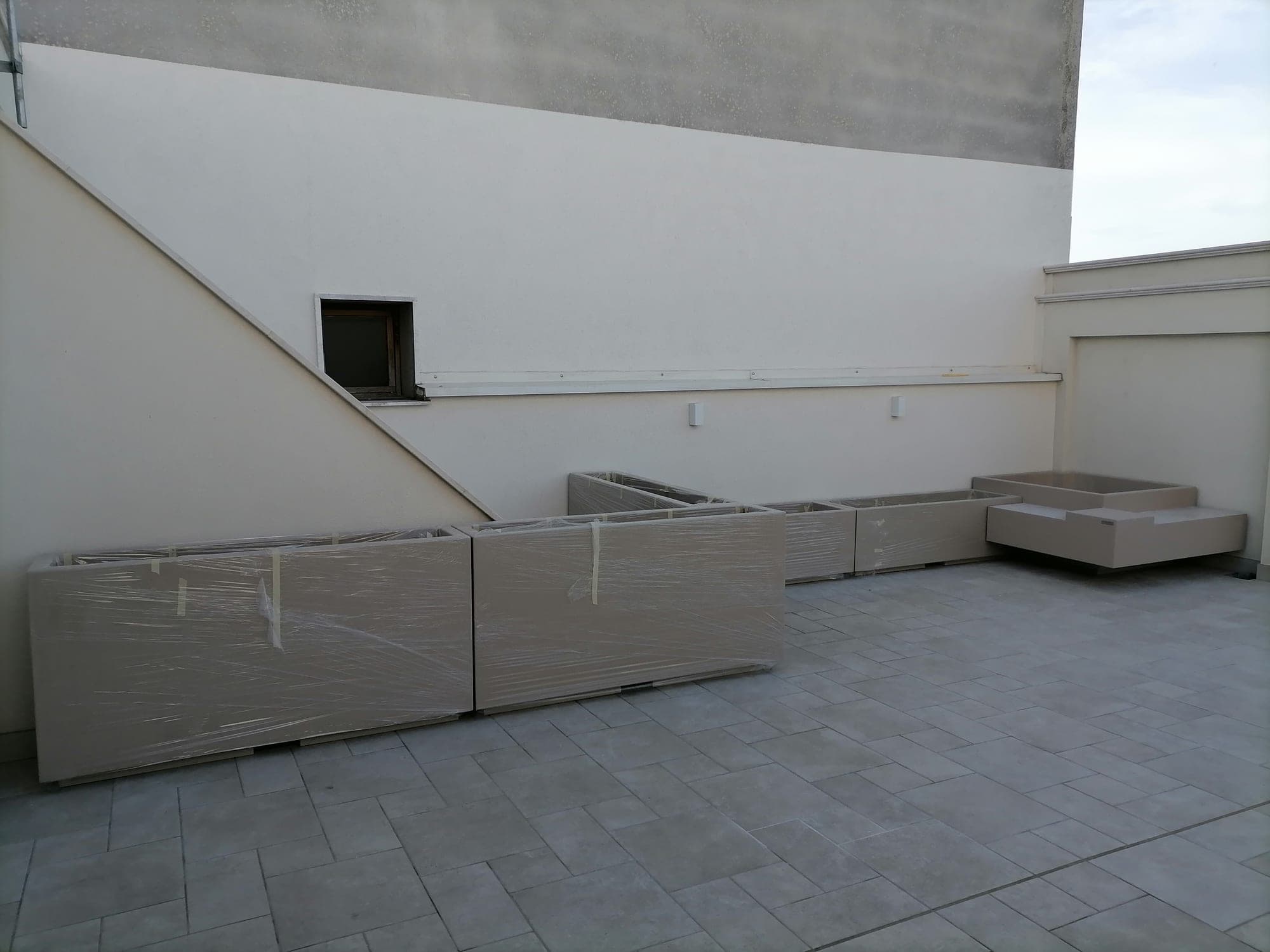 Fioriere color sabbia ancora imballate su un terrazzo, con una seduta ad angolo sullo sfondo