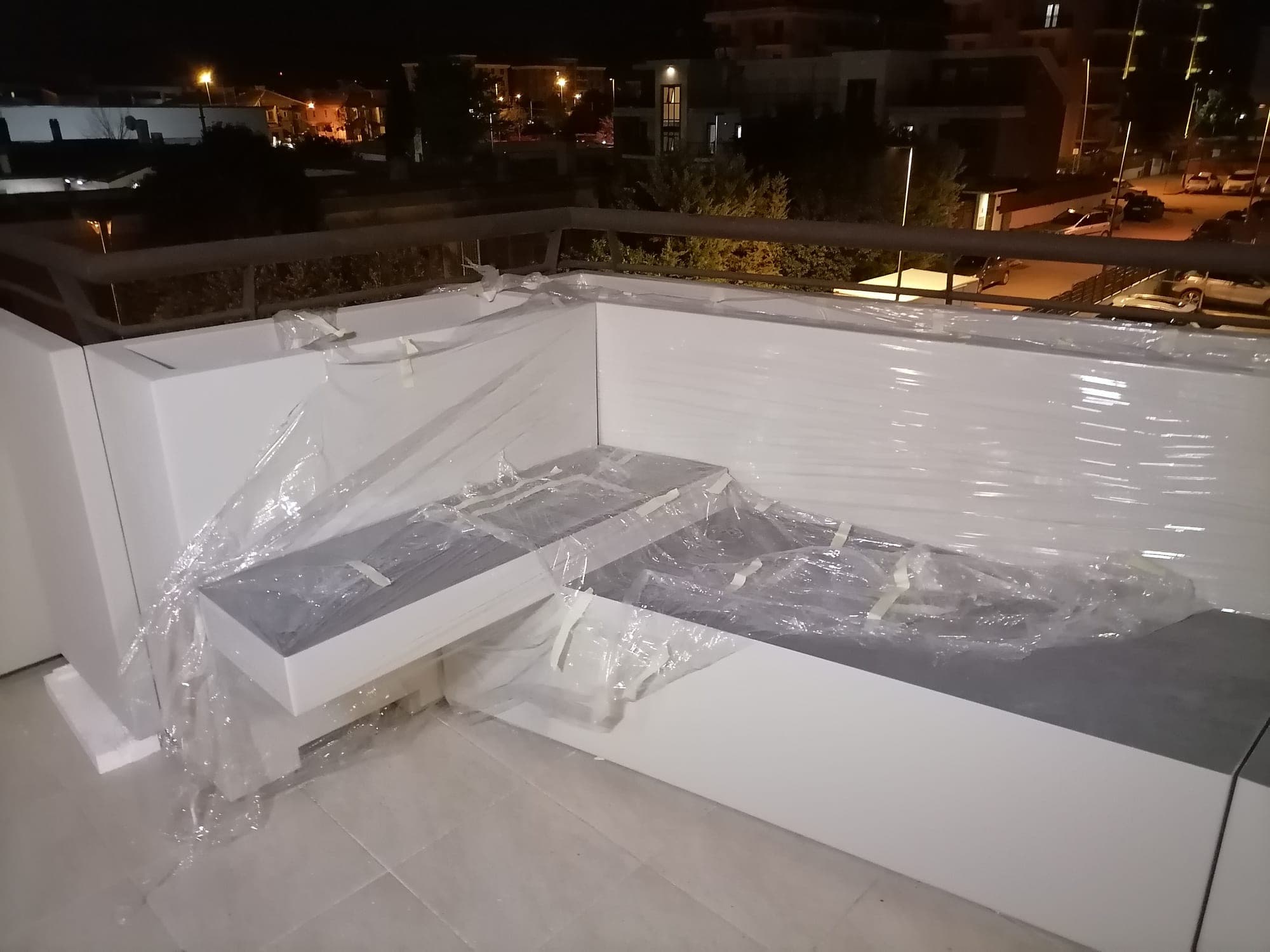 Seduta angolare bianca con piano grigio, ancora avvolta nella pellicola, su un balcone di sera